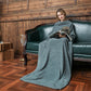 Woman sitting on a green couch reading a book, covered by a gray blanket in a wooden room.