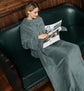 Woman sitting on a green leather couch reading a magazine while wrapped in a gray blanket.