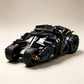 A black and gray toy vehicle built from interlocking blocks, shown on a plain beige studio background. The model has large treaded wheels, a low angular body, and small gold-colored mechanical accents.