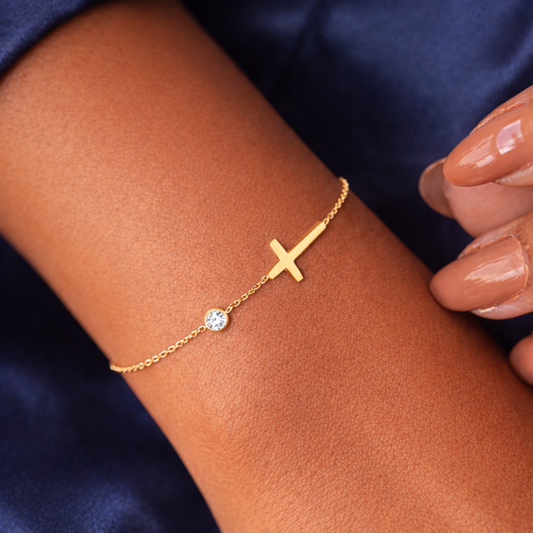 Gold bracelet with a cross and small round charm on a wrist against a dark background