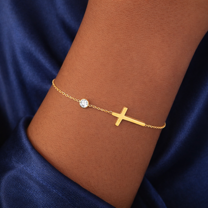 Gold bracelet with a cross and small round element on a wrist against a blue fabric background