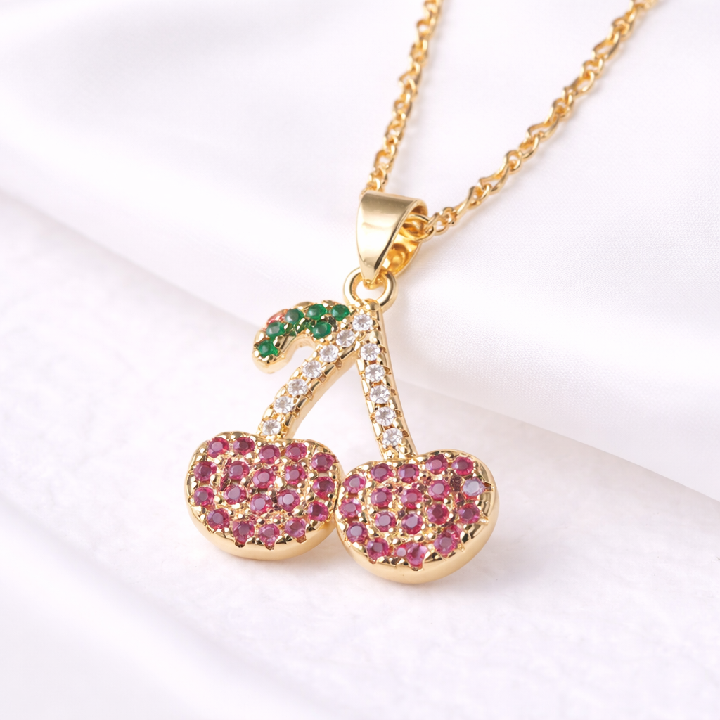 Gold necklace with cherry-shaped pendant featuring pink and green stones on a white background