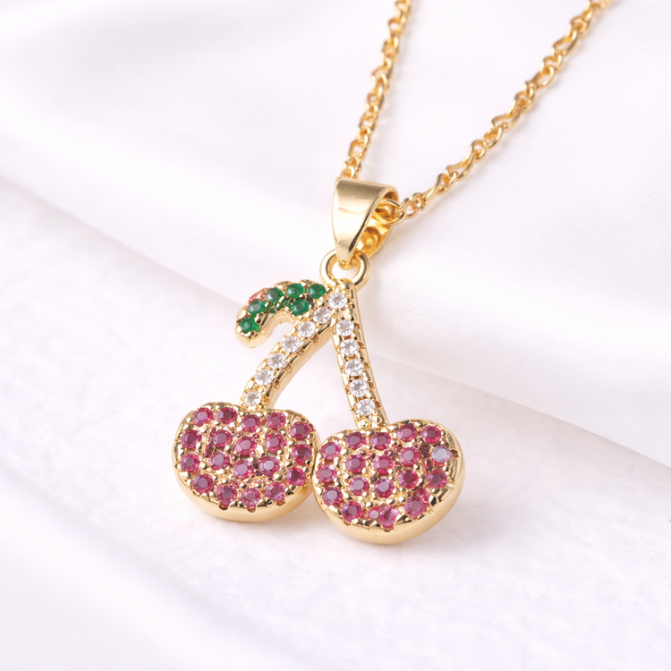 Gold necklace with cherry-shaped pendant featuring pink and green stones on a white background
