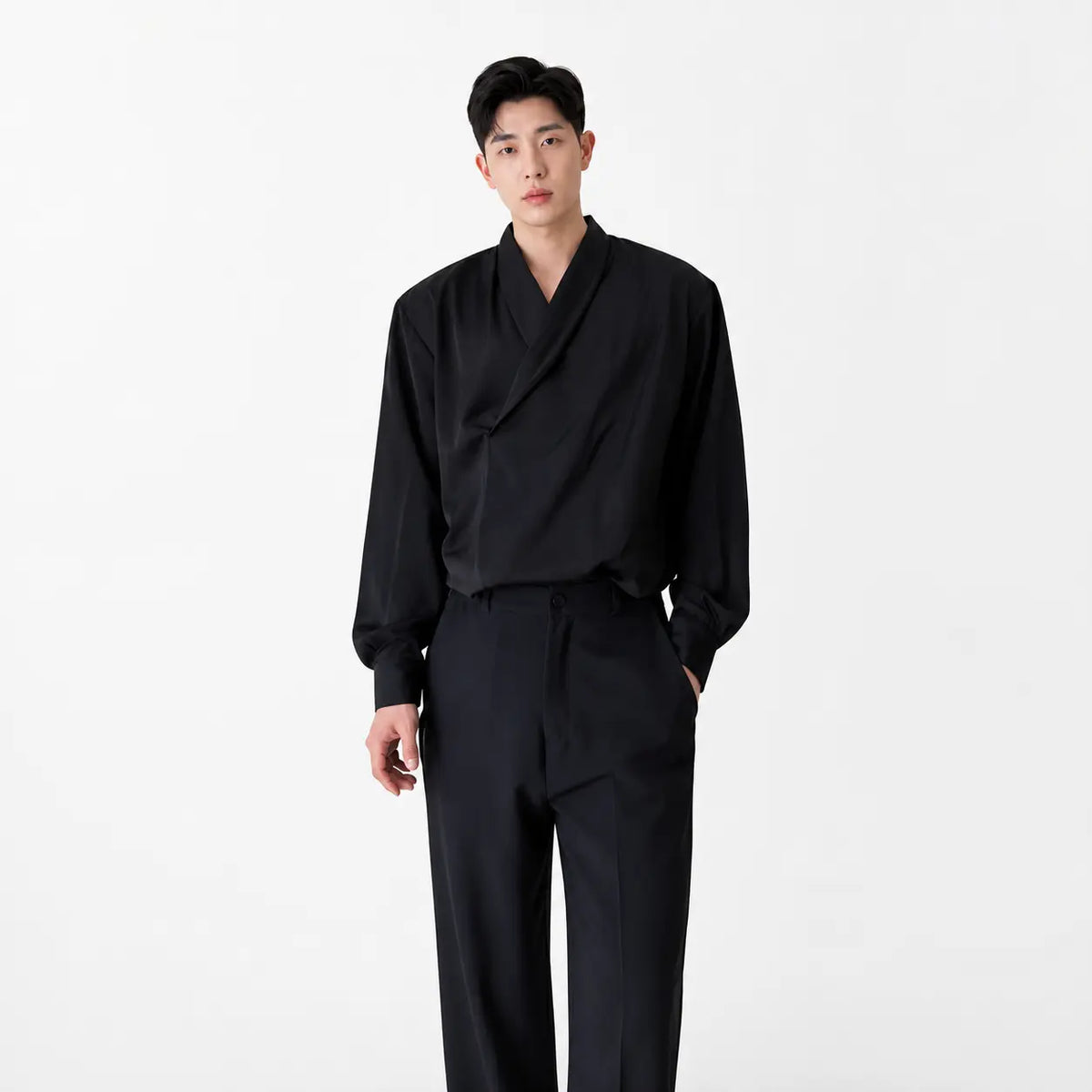 Men's loose fit silk-look shirt in black worn by a model standing with one hand in pocket against white background
