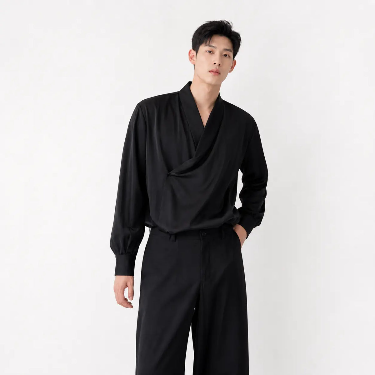Men's loose fit silk-look shirt in black worn by a model with hands in pockets against white background