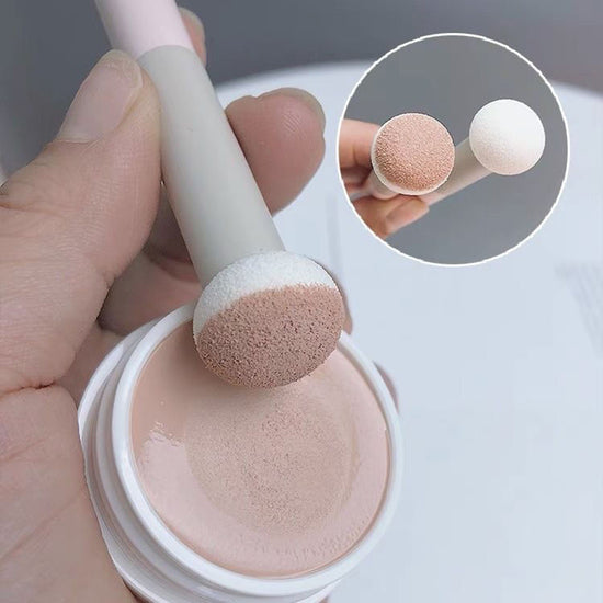 Makeup brush with pink sponge tip being dipped into a container of beige cream, with a close-up inset showing the brush tip.