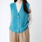 Person wearing a turquoise knitted vest over a white shirt and beige pants on a white background