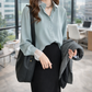 Woman in a light blue shirt and black skirt holding a black bag and gray coat in an office setting.