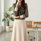 Woman in a patterned blouse and beige skirt standing in an office setting with a desk and plant.