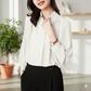 Woman wearing a white blouse and black pants in a bright room with a plant and shelves.