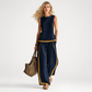 Woman wearing a navy outfit with gold trim holding a woven bag on a white background
